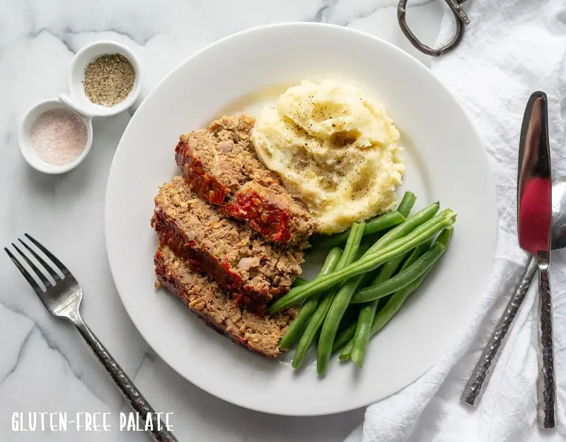 sliced Gluten-Free Meatloaf on a white plate with mashed potatoes and green beans, next to a fork and knife