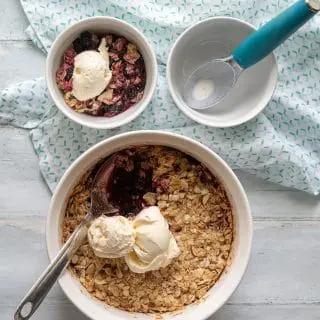 a close up of blackberry crisp topped with two scoops of vanilla ice cream, next to a bowl of blackberry crisp and a ice cream scoop
