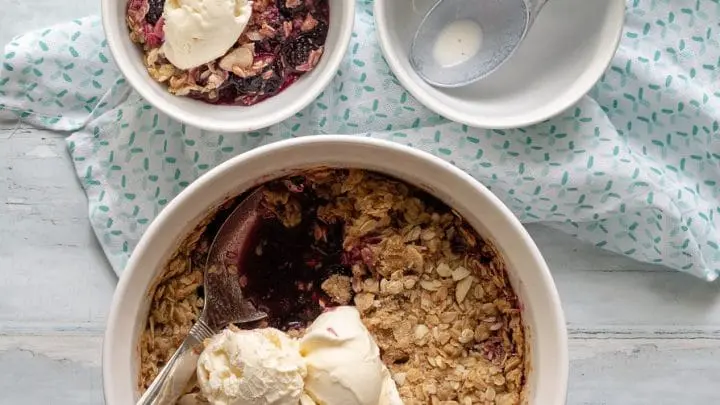 a close up of blackberry crisp topped with two scoops of vanilla ice cream, next to a bowl of blackberry crisp and a ice cream scoop