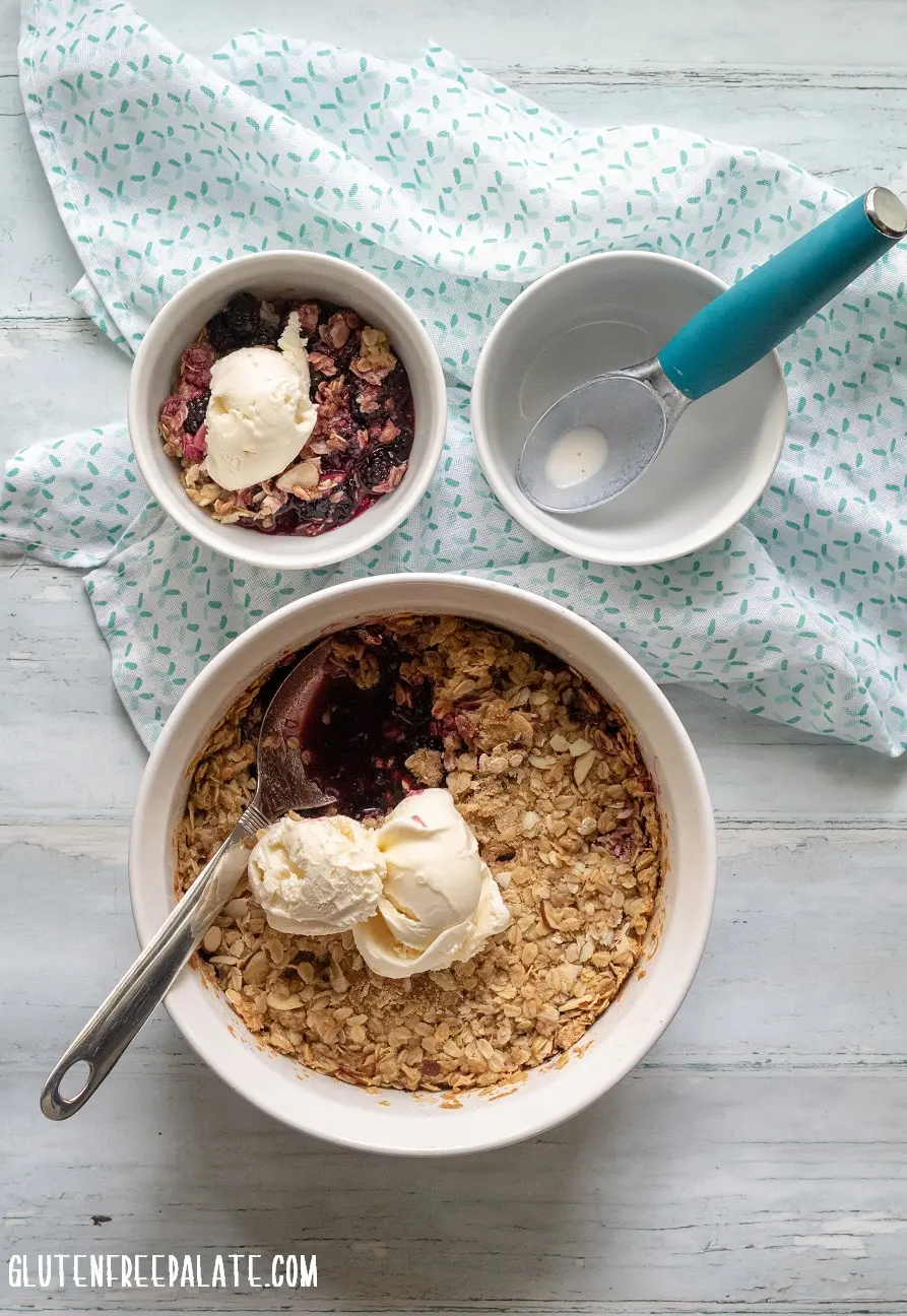 blackberry crisp topped with two scoops of vanilla ice cream, next to a bowl of blackberry crisp and a ice cream scoop