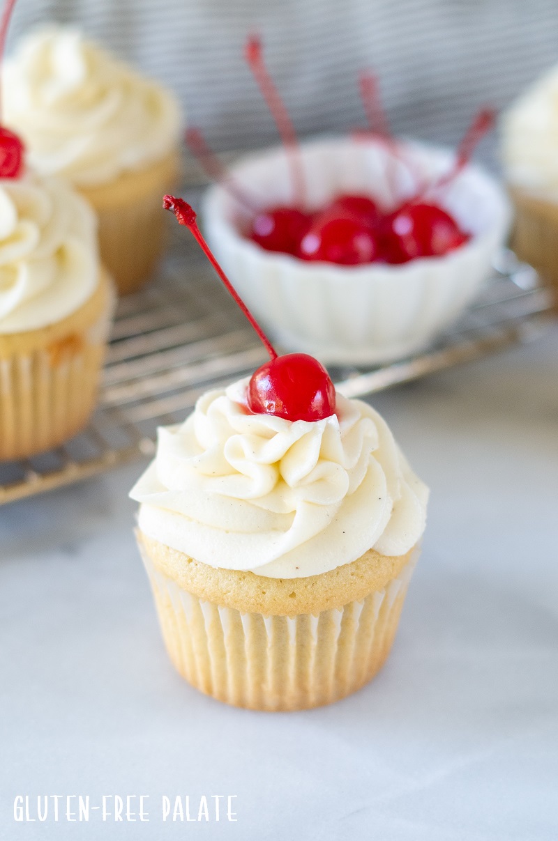 a white Gluten Free Root Beer Cupcake topped with white frosting with a cherry on top and a bowl of red cherries in the background