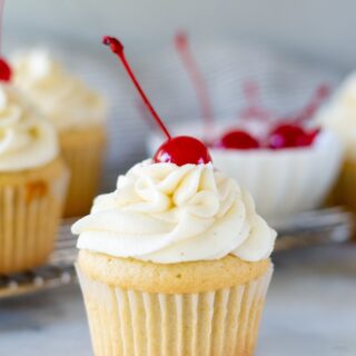 a white Gluten Free Root Beer Cupcake topped with white frosting with a cherry on top and a bowl of red cherries in the background