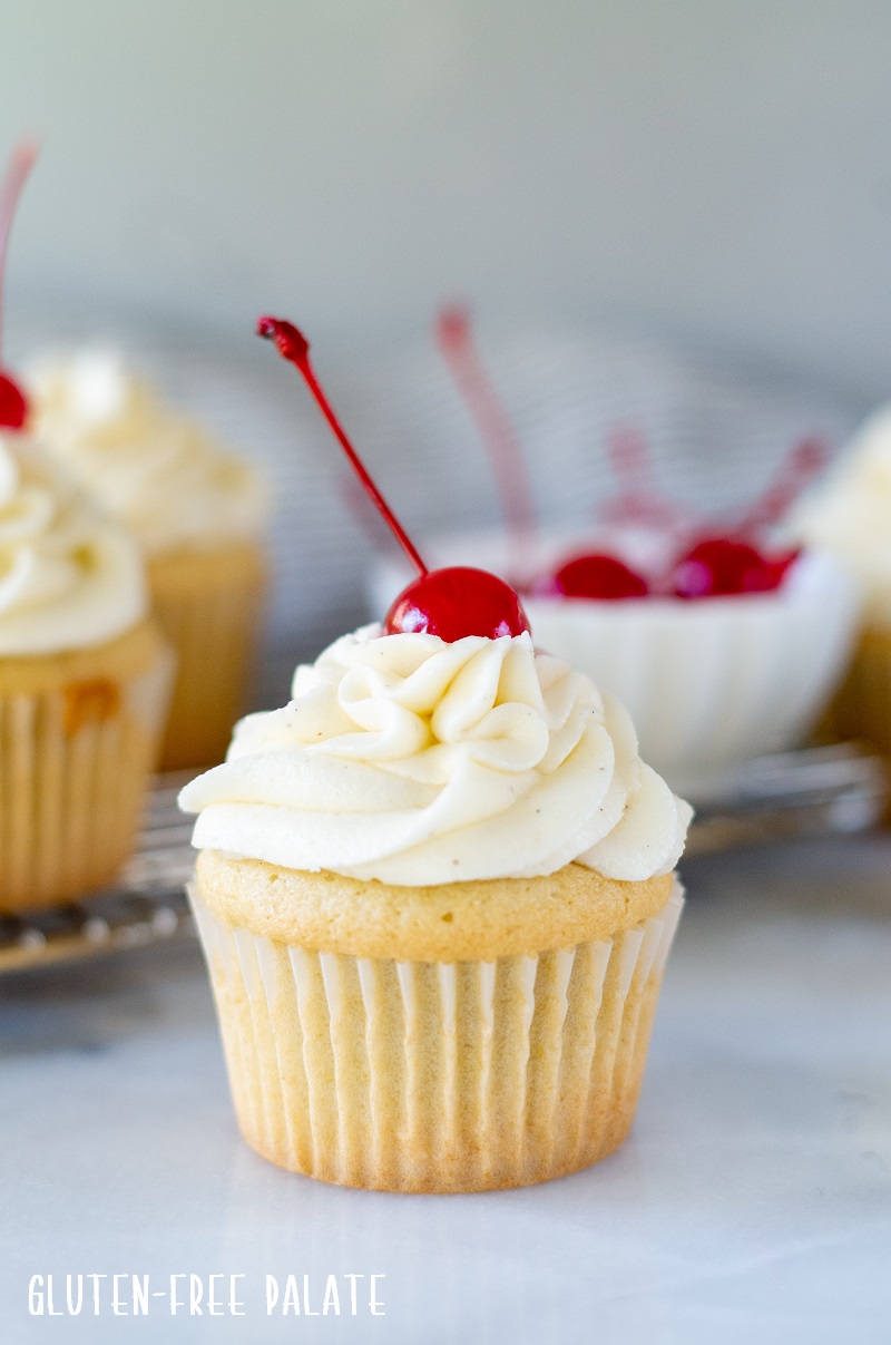 close up of a white Gluten Free Root Beer Cupcake topped with white frosting with a cherry on top and a bowl of red cherries in the background