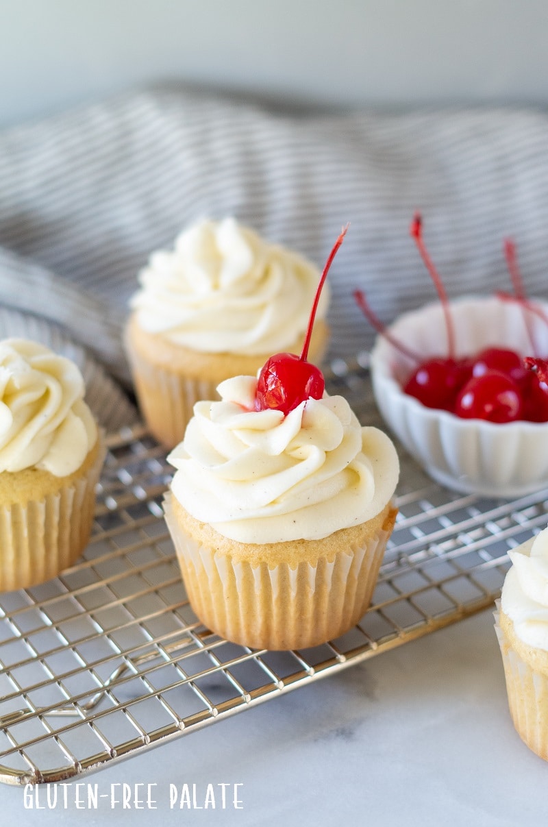 a white Gluten Free Root Beer Cupcake topped with white frosting with a cherry on top and a bowl of red cherries and additional cupcakes in the background