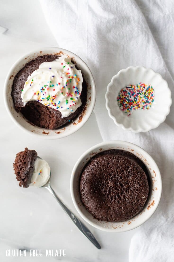 gluten free mug cake in a white ramekin with whipped cream next to a ramekin with cake and a spoon with a bite out