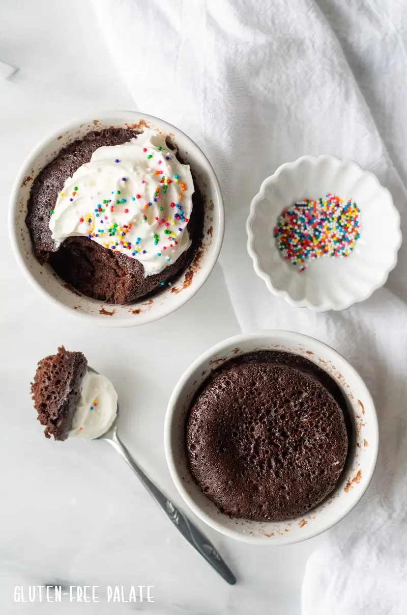 gluten-free chocolate mug cake in a white ramekin with whipped cream next to a ramekin with cake and a spoon with a bite out