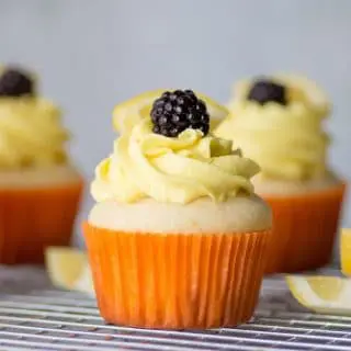 a close up of a gluten-free lemon cupcake topped with yellow frosting and a blackberry, in a orange paper liner