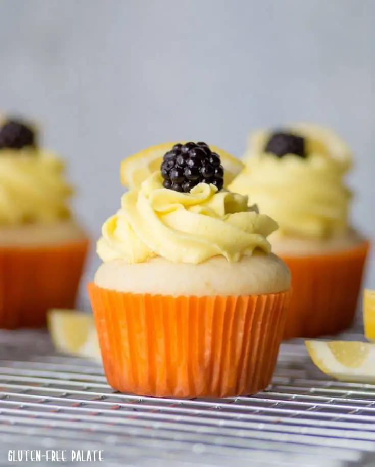 a close-up of a gluten-free lemon cupcake topped with yellow frosting and blackberry, in an orange paper liner