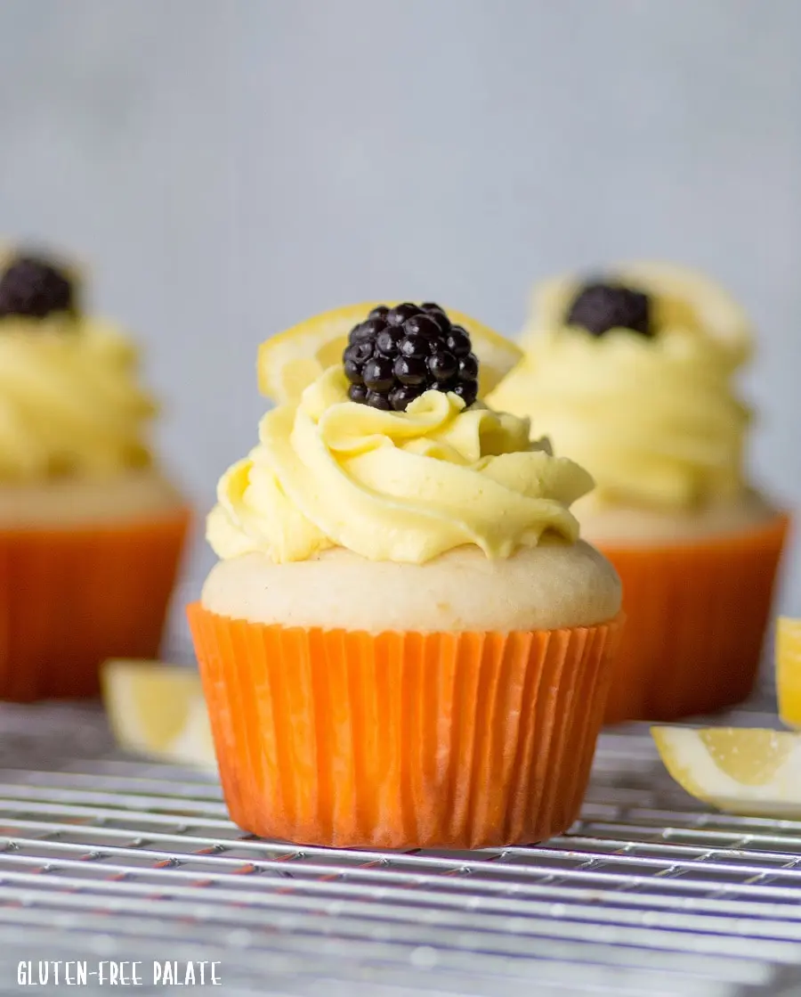 a gluten-free lemon cupcake in an orange paper wrapper, topped with yellow frosting and a blackberry