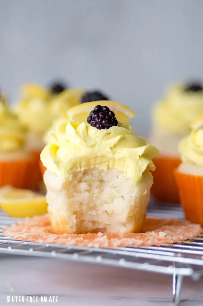 a close up of a lemon cupcake with a bite out topped with yellow frosting and a blackberry, in a orange paper liner