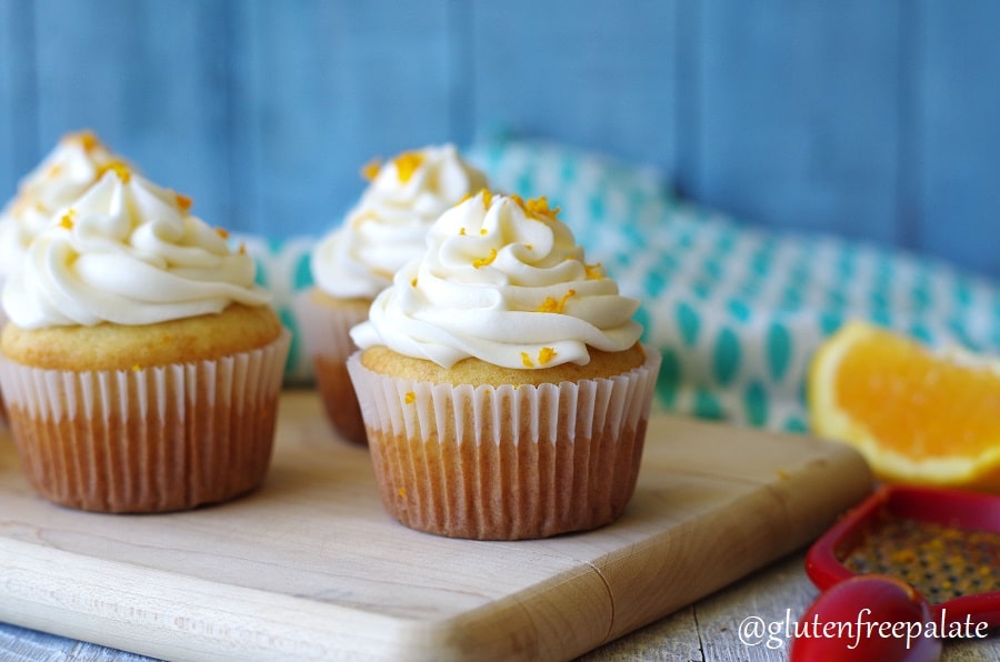three orange cupcakes with white frosting and orange peel on top, on a cutting board next to a orange wedge