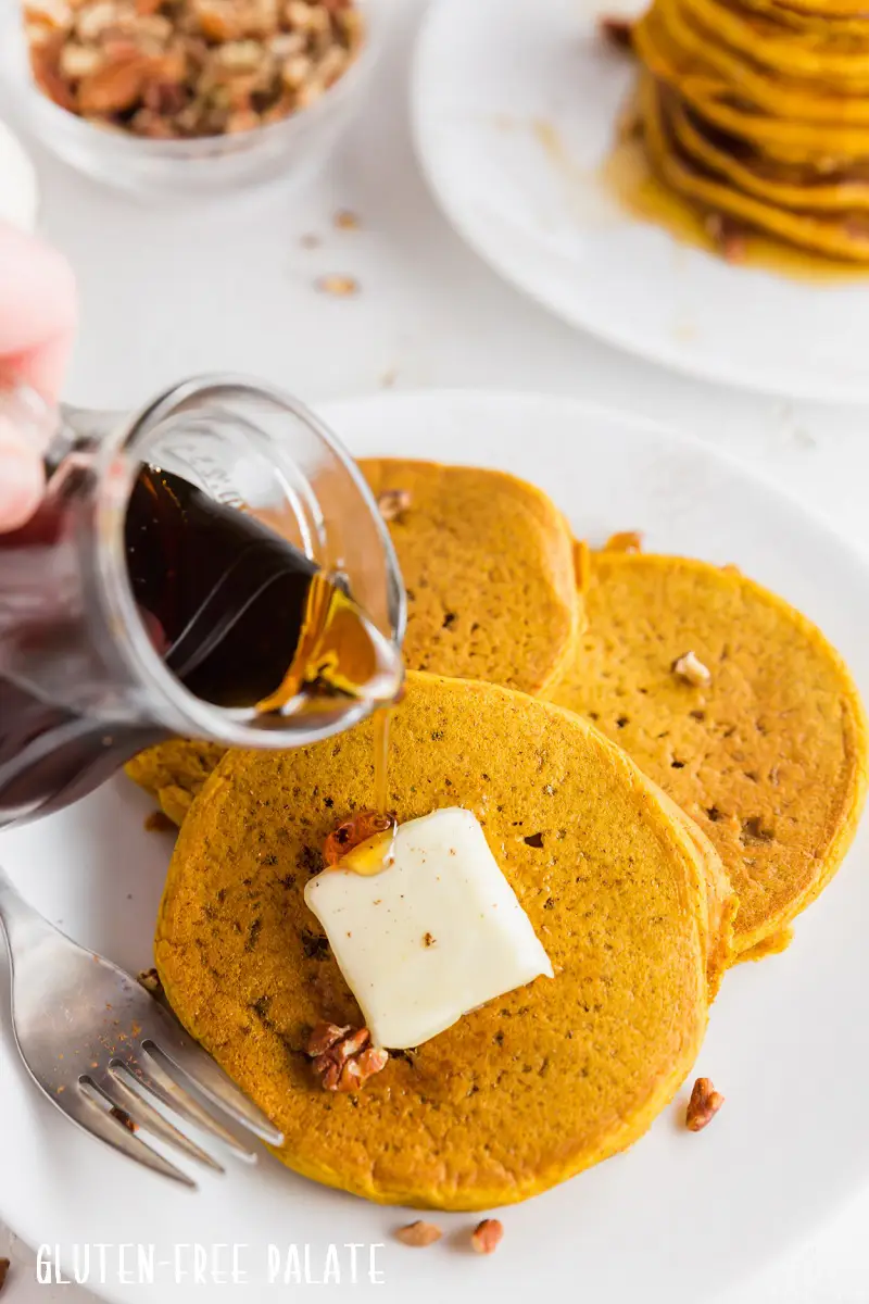 a hand pouring syrup on top of gluten-free pumpkin pancakes.