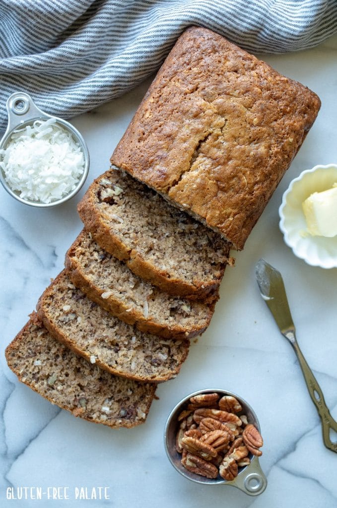 Gluten-Free Banana Bread sliced on marble slab next to a measuring cup of coconut and a measuring cup of pecans
