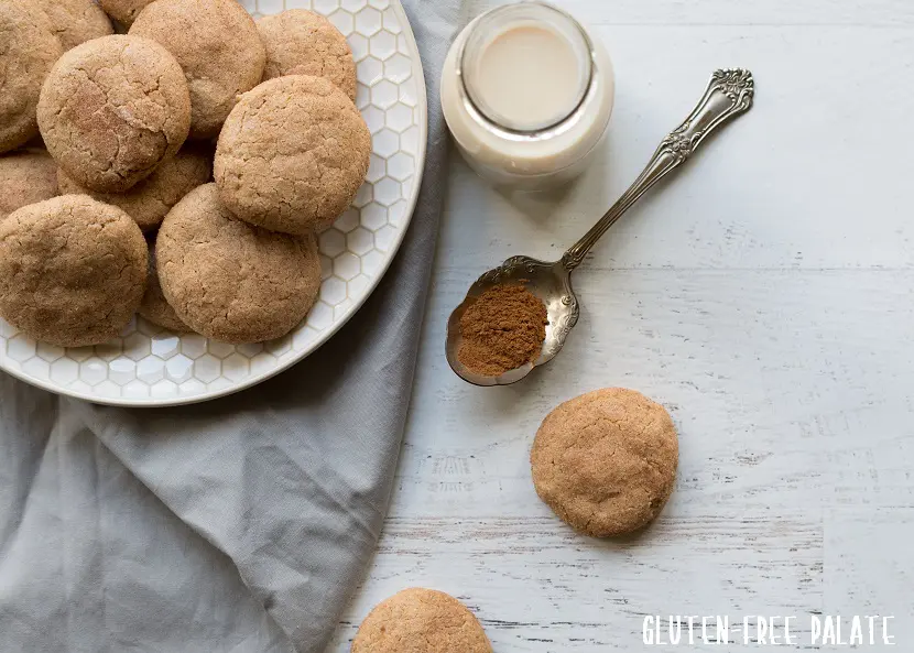 a close up of two paleo snickerdoodle cookies next to a plate of cookies and a spoon of cinnamon