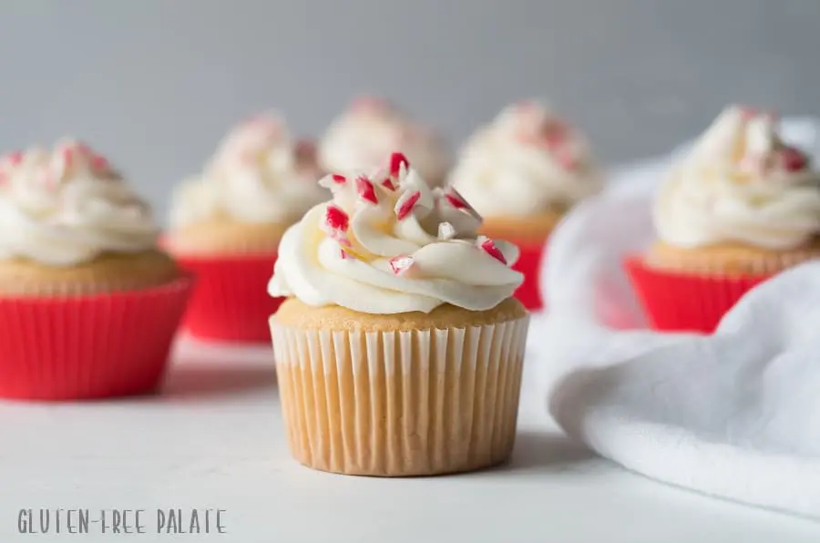 a cupcake topped with white frosting and candy cane pieces set in front of other cupcakes with red paper liners