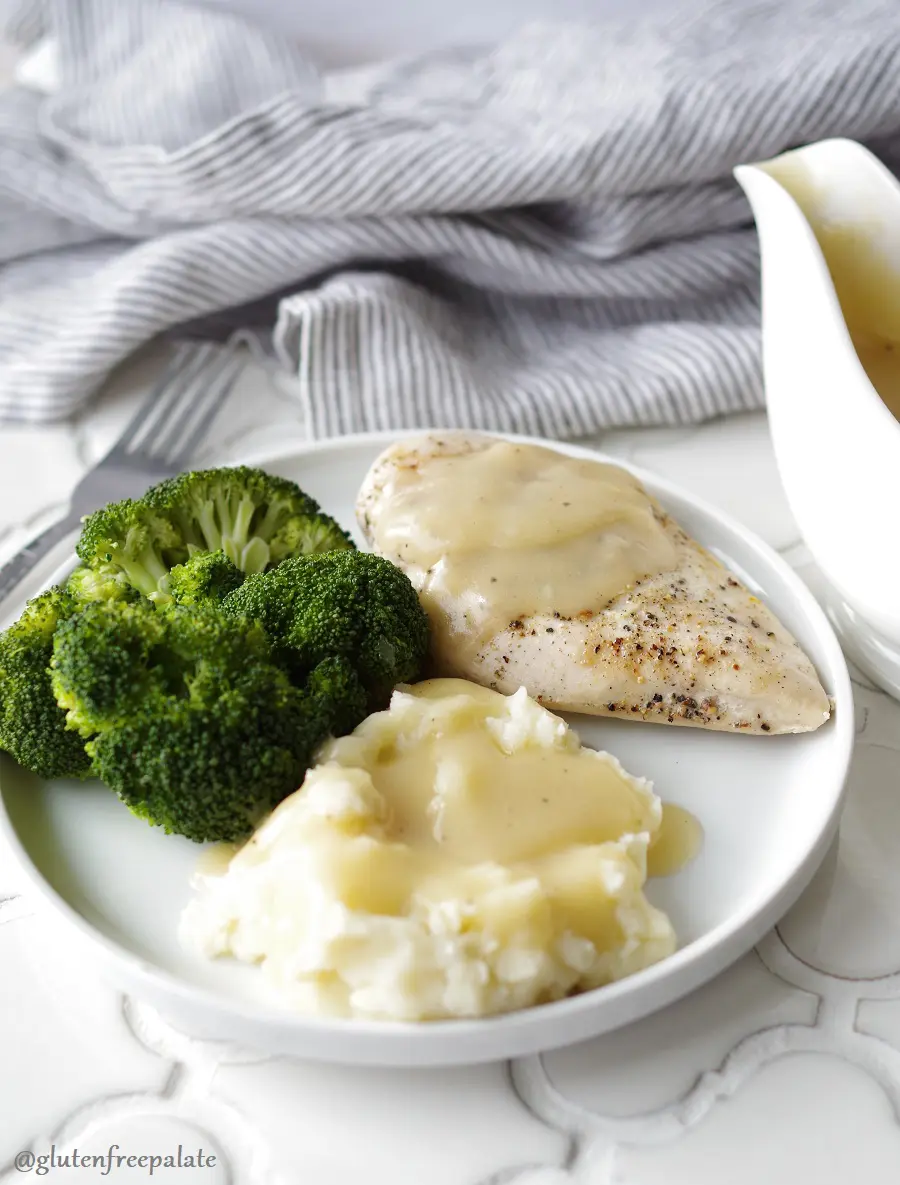 mashed potatoes and gravy, broccoli, and chicken with gravy on a white plate next to a gravy boar and a stripe towel