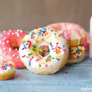 a close up of gluten free vanilla donuts with colored icing and colored sprinkles arranged next to a jar of milk