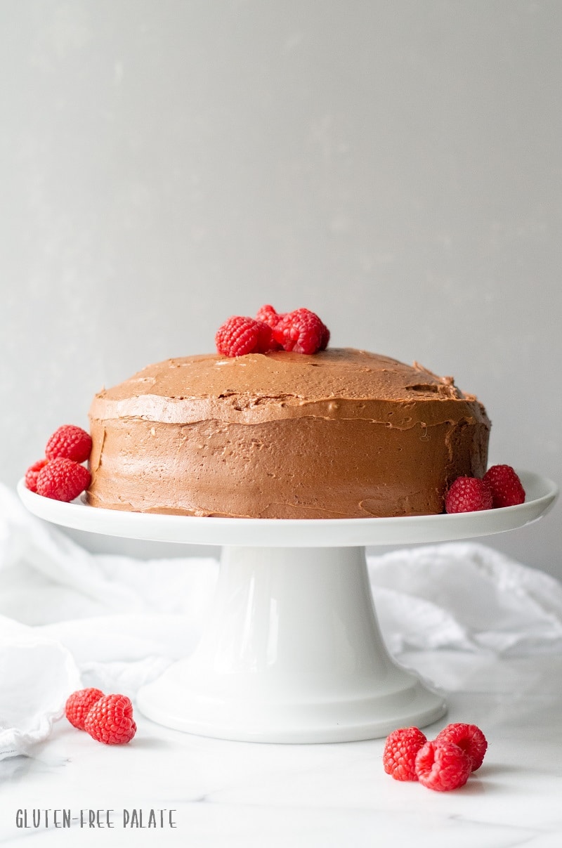 a straight-on view of a chocolate cake topped with chocolate frosting and raspberries on a white cake stand