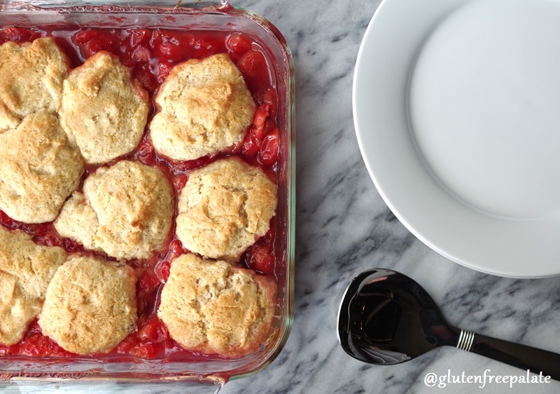 Gluten free cherry cobbler in a squre baking dish next to a white plate and a spoon