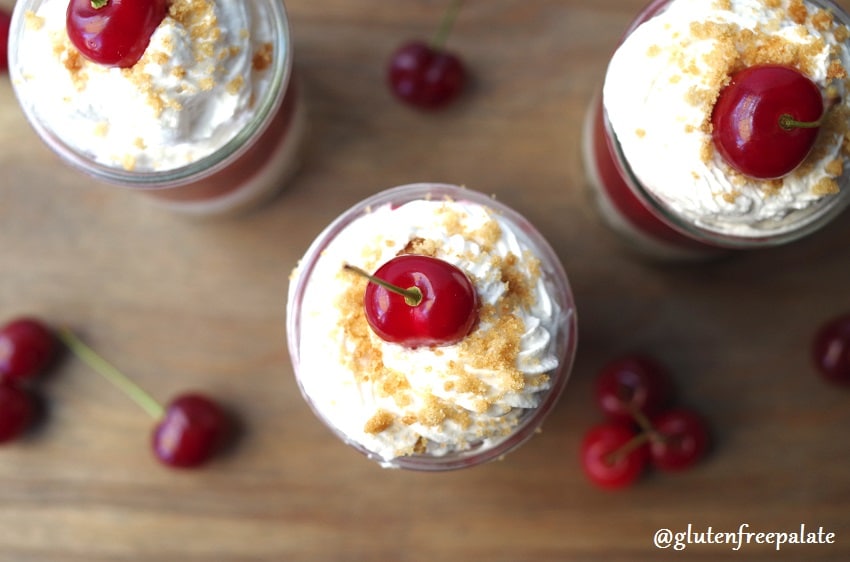 a top down view of three shot glasses filled with layers of graham cracker crumbs, whipped cream, cherry pie filling, topped with whipped cream and a cherry