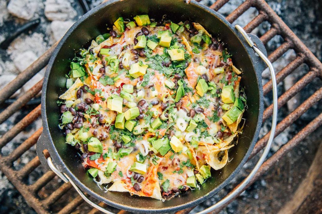 a cast iron pot filled with nacho's topped with avocado pieces, over a fire