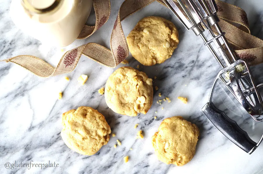 four gluten-free white chocolate macadamia cookies on a marble slab with a browni ribbon and hand beaters