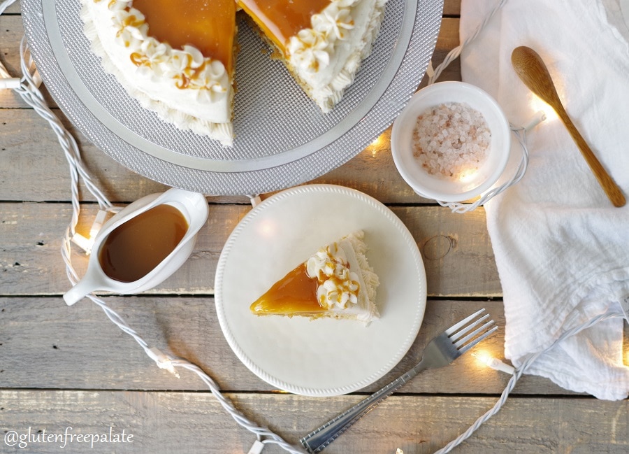 top down view of a slice of gluten free caramel cake topped with frosting and caramel next to lights, a fork, salt, and a wooden spoon