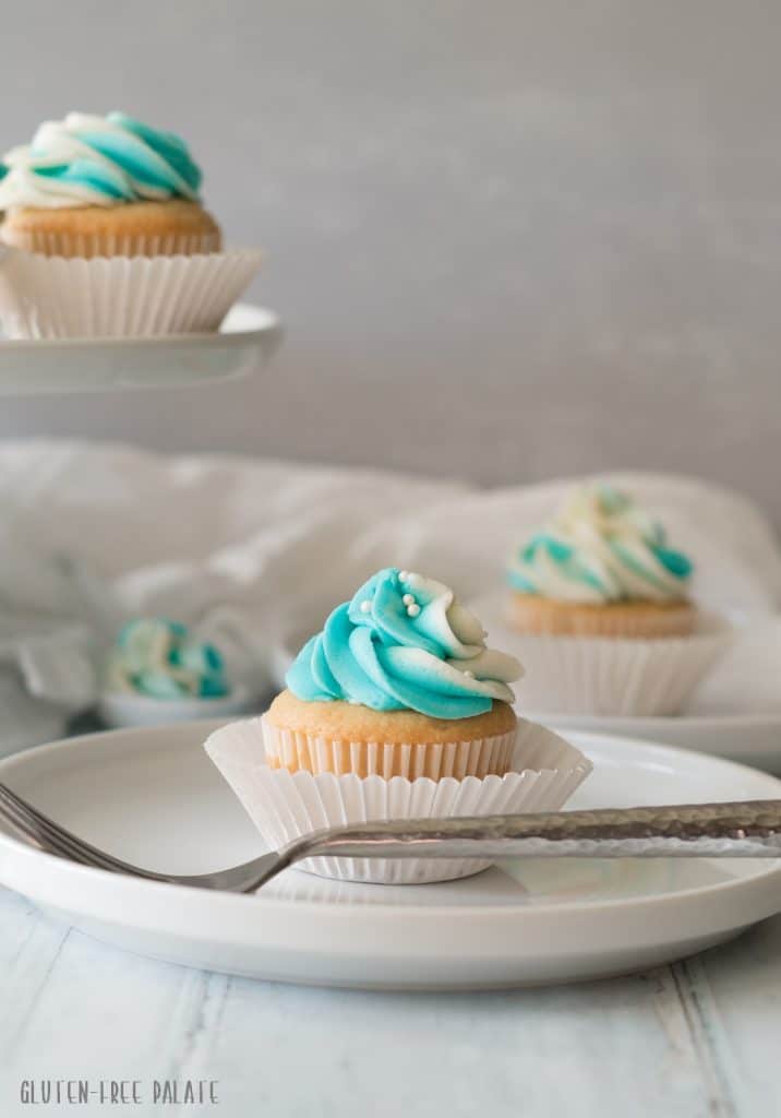 a vanilla cupcake topped with blue and white swirled frosting in a white paper liner, on a white plate