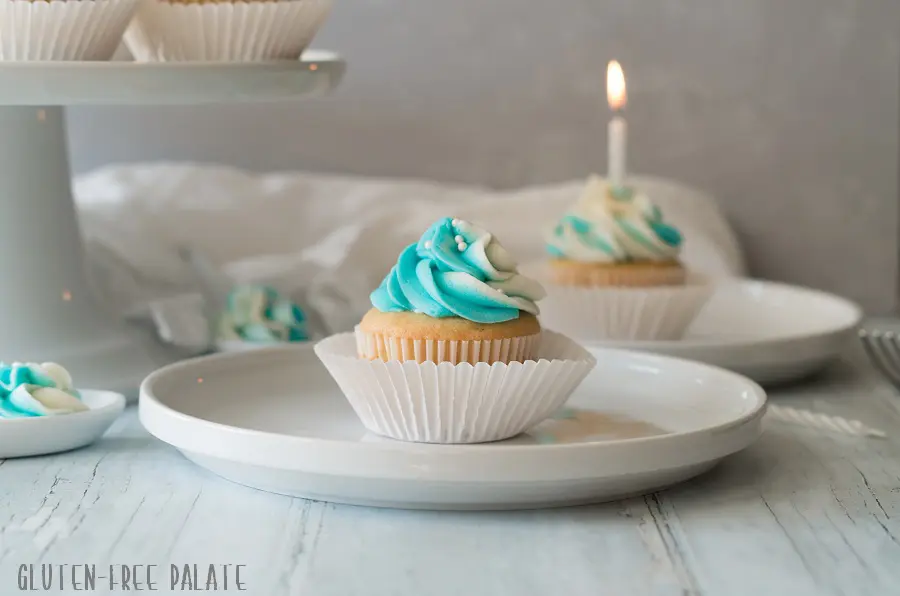 a gluten free vanilla cupcake topped with blue and white swirled frosting, on a white plate in front of another cupcake