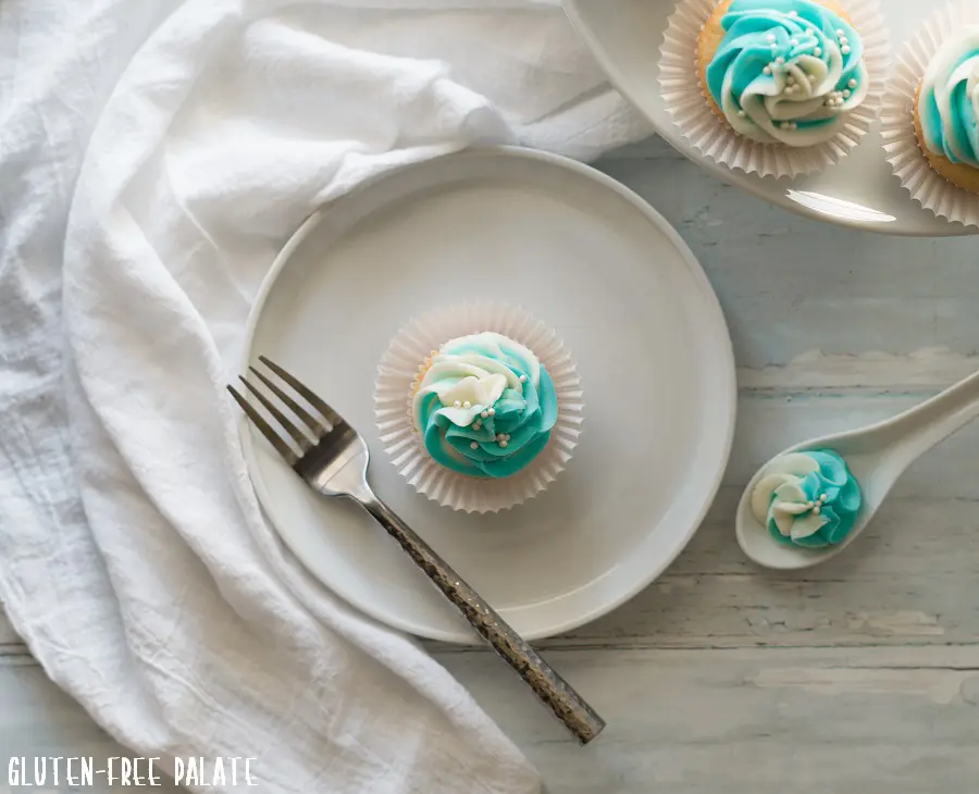top down view of a gluten free vanilla cupcake topped with blue and white swirled frosting, on a white plate with a fork