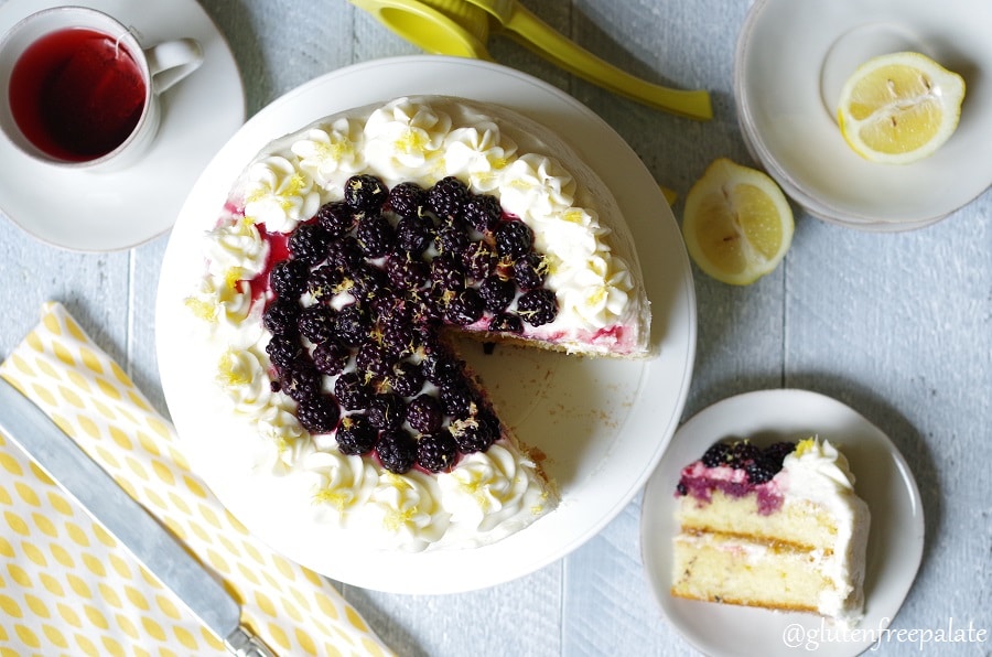 top down view of a lemon cake topped with white frosting and blackberries, with a slice of cake on a white plate next to it, lemon wedges, tea and a yellow napkin