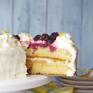 a close up of a serving spoon serving a slice of gluten-free lemon cake topped with white frosting and blackberries