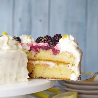 a close up of a serving spoon serving a slice of gluten-free lemon cake topped with white frosting and blackberries