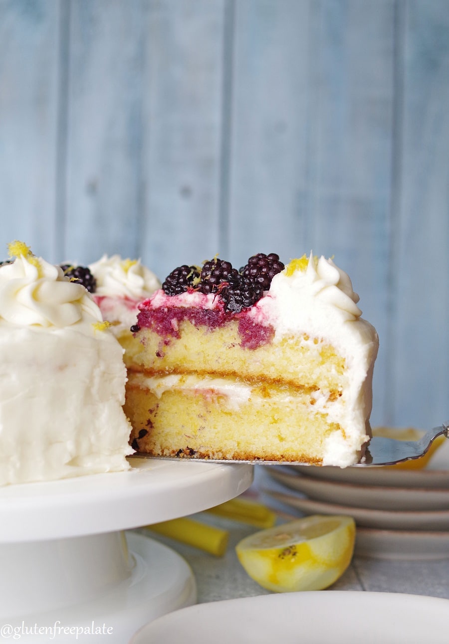 serving knife with a slice of lemon cake on it on a white cake plate with lemon and blackberries
