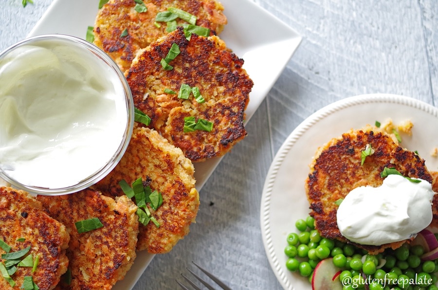 Gluten-Free Salmon Cakes on a white plate with green garnish, and a bowl of white sauce, next to a plate of salmon cakes and peas