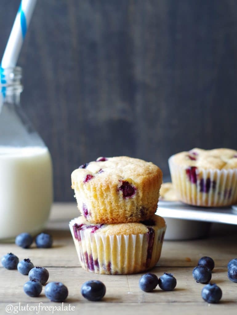 two gluten free blueberry muffins stacked next to a jar of milk and scattered blueberries