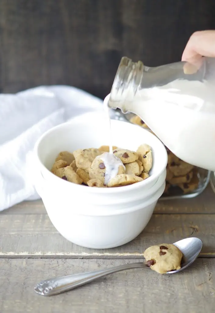 milk being poured over gluten free vegan cereal in the shape of chocolate chips cookies in a white bowl