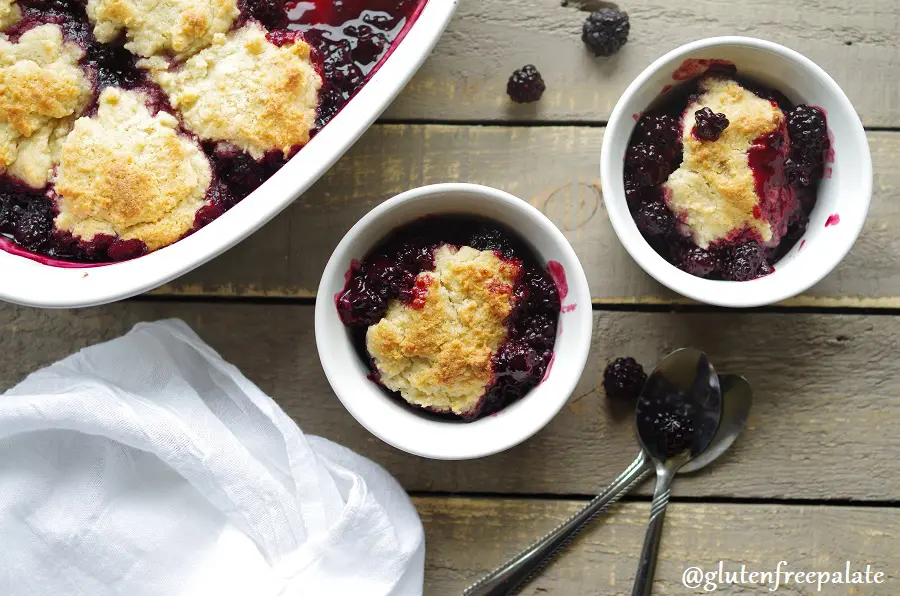 Gluten-Free Blackberry Cobbler in a white baking dish next to two white bowls of blackberry cobbler, and two spoons