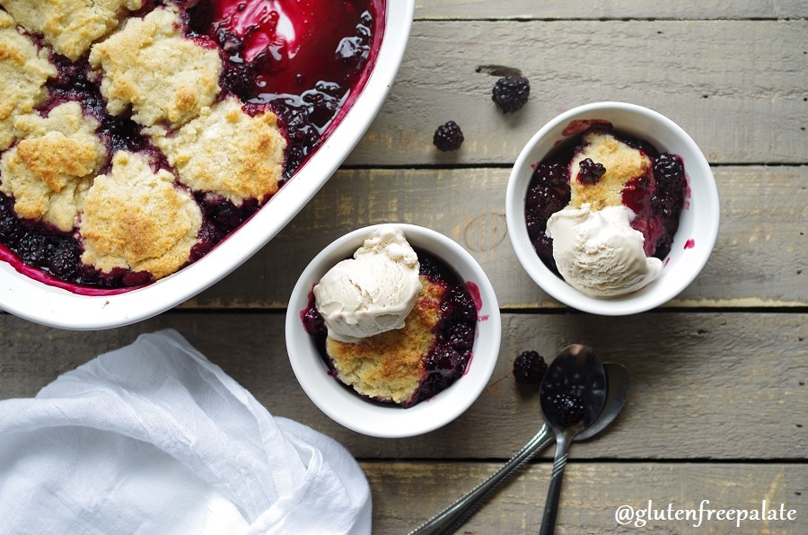 a top down view of Gluten-Free Blackberry Cobbler in white bowls with vanilla ice cream next to a baking dish filled with cobbler