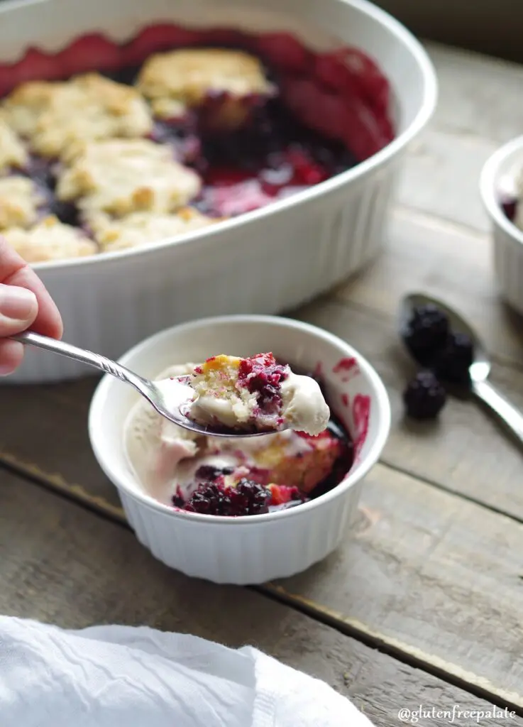 a spoon with a bite of Gluten-Free Blackberry Cobbler next to a white bowl filled blackberry cobberl and vanilla ice cream.
