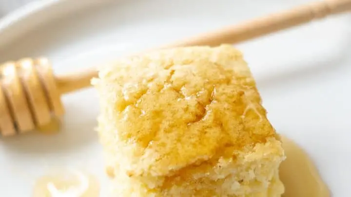 A slice of gluten-free cornbread on a white place with a honey drizzle.