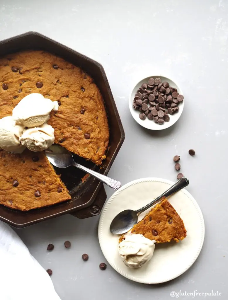 gluten free pumpkin chocolate chip skillet cookie in a cast iron skillet topped with ice cream next to a piece of cookie on a white plate