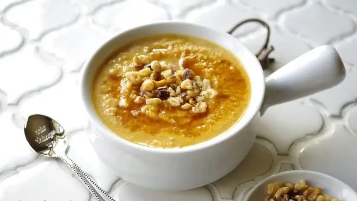 a side view of pumpkin porridge in a white bowl topped with chopped nuts, next to a spoon