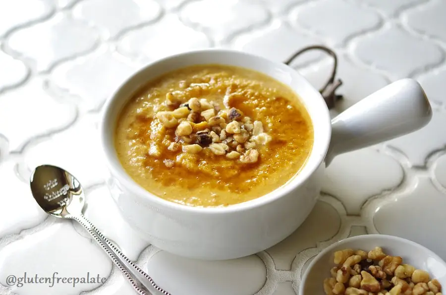 a side view of pumpkin porridge in a white bowl topped with chopped nuts, next to a spoon and a bowl of chopped nuts