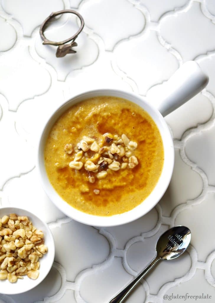 a top down view of pumpkin porridge in a white bowl topped with chopped nuts, next to a spoon a a bowl of chopped nuts