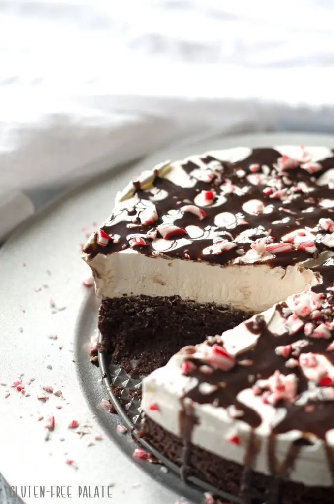  a side view of a brownie ice cream cake topped with chocolate sauce and crushed candy cane pieces, with a slice cut out so you can see the center