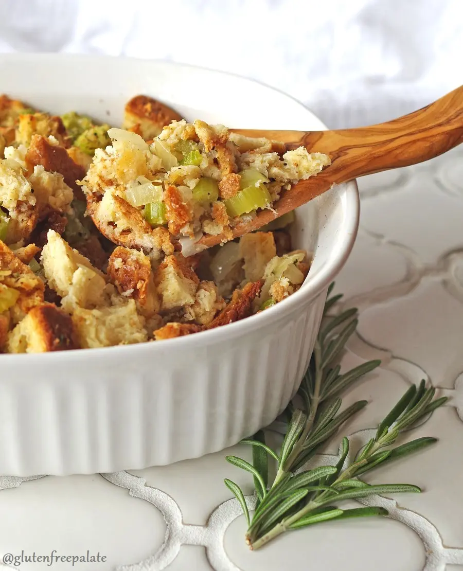 a white baking dish with gluten free stuffing bread cubes, celery and onion with a wood spoon dipped in