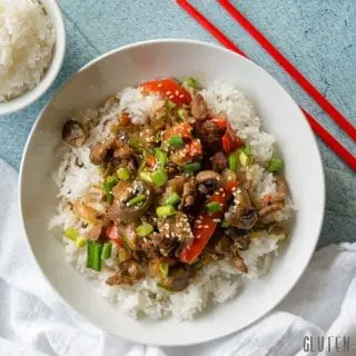 a close up of mongolian beef and vegetables over rice in a white bowl, red chopstick are resting on the side of the bowl