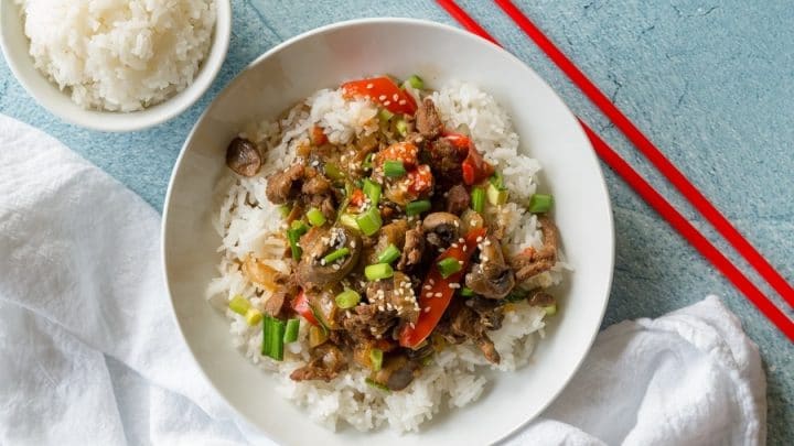 a close up of mongolian beef and vegetables over rice in a white bowl, red chopstick are resting on the side of the bowl