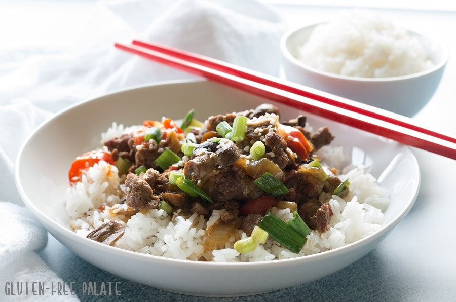 a side view of mongolian beef and vegetables over rice in a white bowl, red chopstick are resting on the side of the bowl